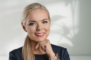 Edyta Kozera. Blondynka o długich włosach, ubrana w ciemną bluzkę, uśmiecha się do zdjęcia na białym tle.